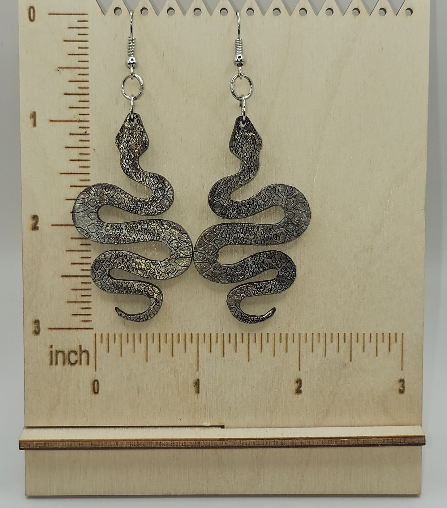 Silver snake-shaped earrings on a wooden display card with a ruler.
