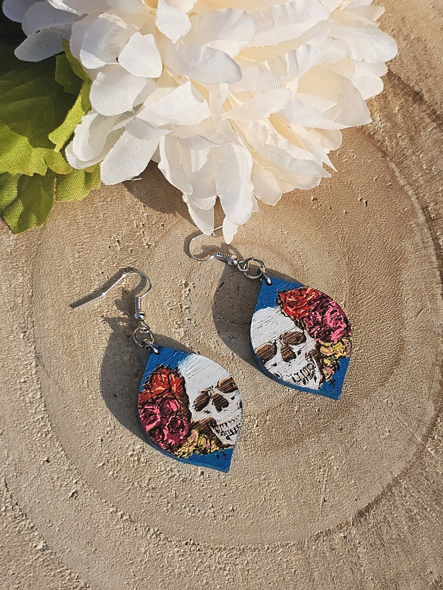 Pair of earrings with skull and floral design on a wood surface with flowers in the background
