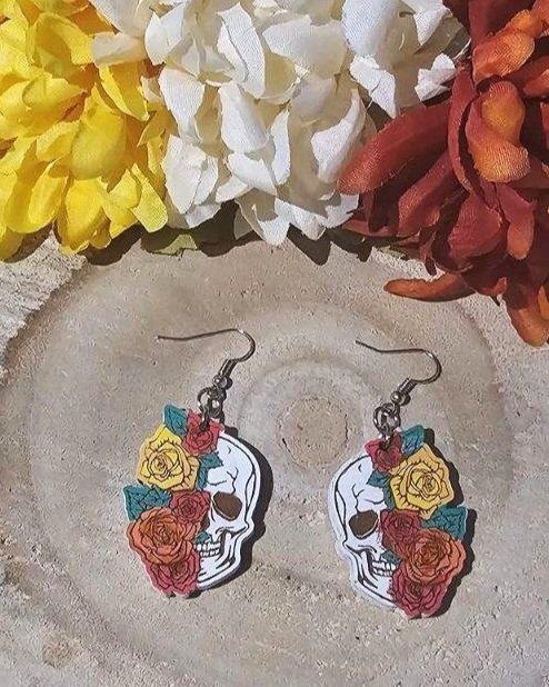 Skull-shaped earrings with floral designs on a wood surface with flowers in the background
